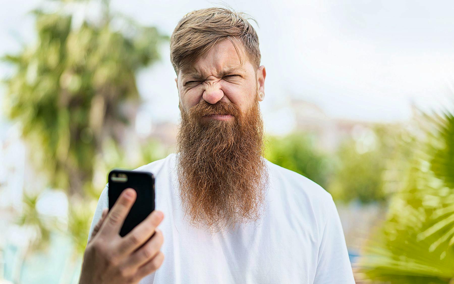 Ein junger Mann mit langem Bart verzieht das Gesicht und schaut genervt auf sein Smartphone.