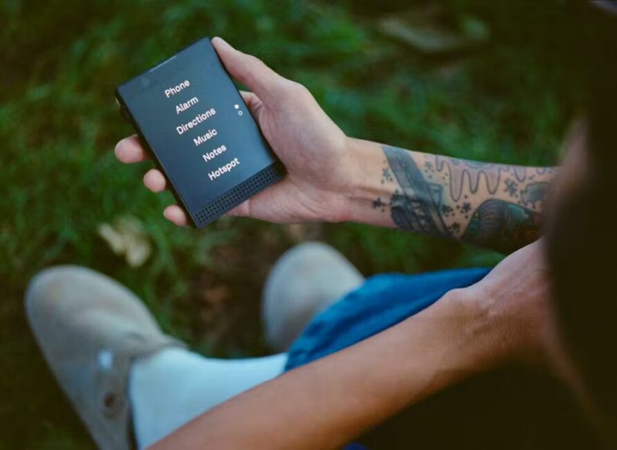 Persona tatuata tiene in mano uno smartphone semplice con scritte grandi, seduta sull’erba.