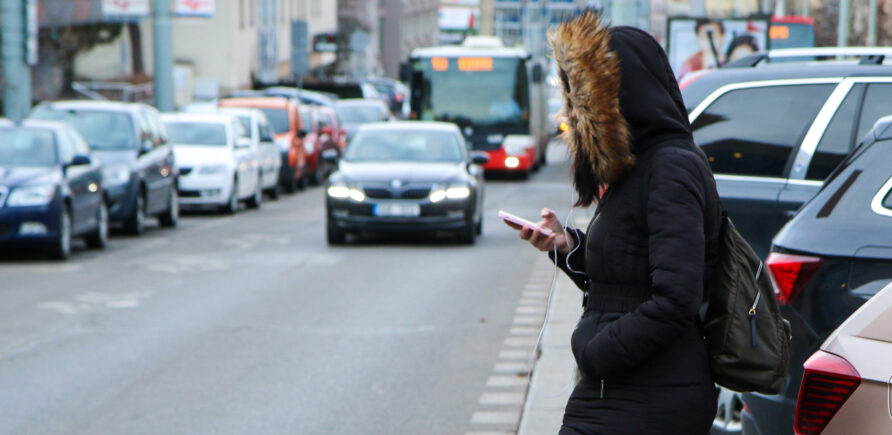 Une personne avec une veste d’hiver et une capuche traverse sans faire attention une route très fréquentée tout en regardant son smartphone.