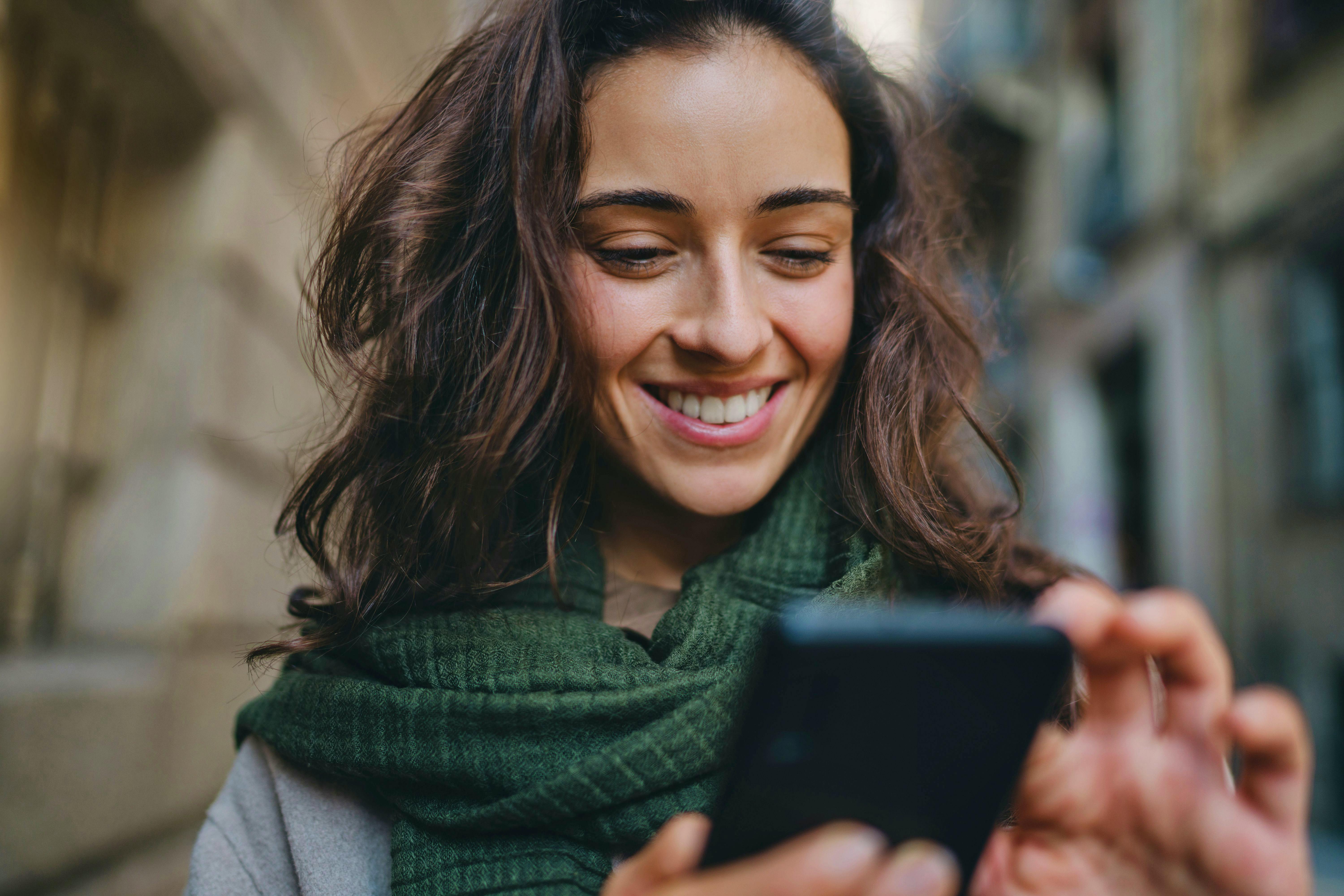 Una giovane donna sorride mentre guarda il telefono per strada.