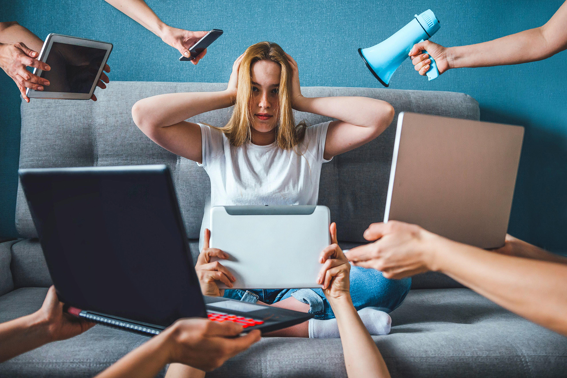 Una giovane donna stressata sul divano si tappa le orecchie, circondata da dispositivi digitali e un megafono.