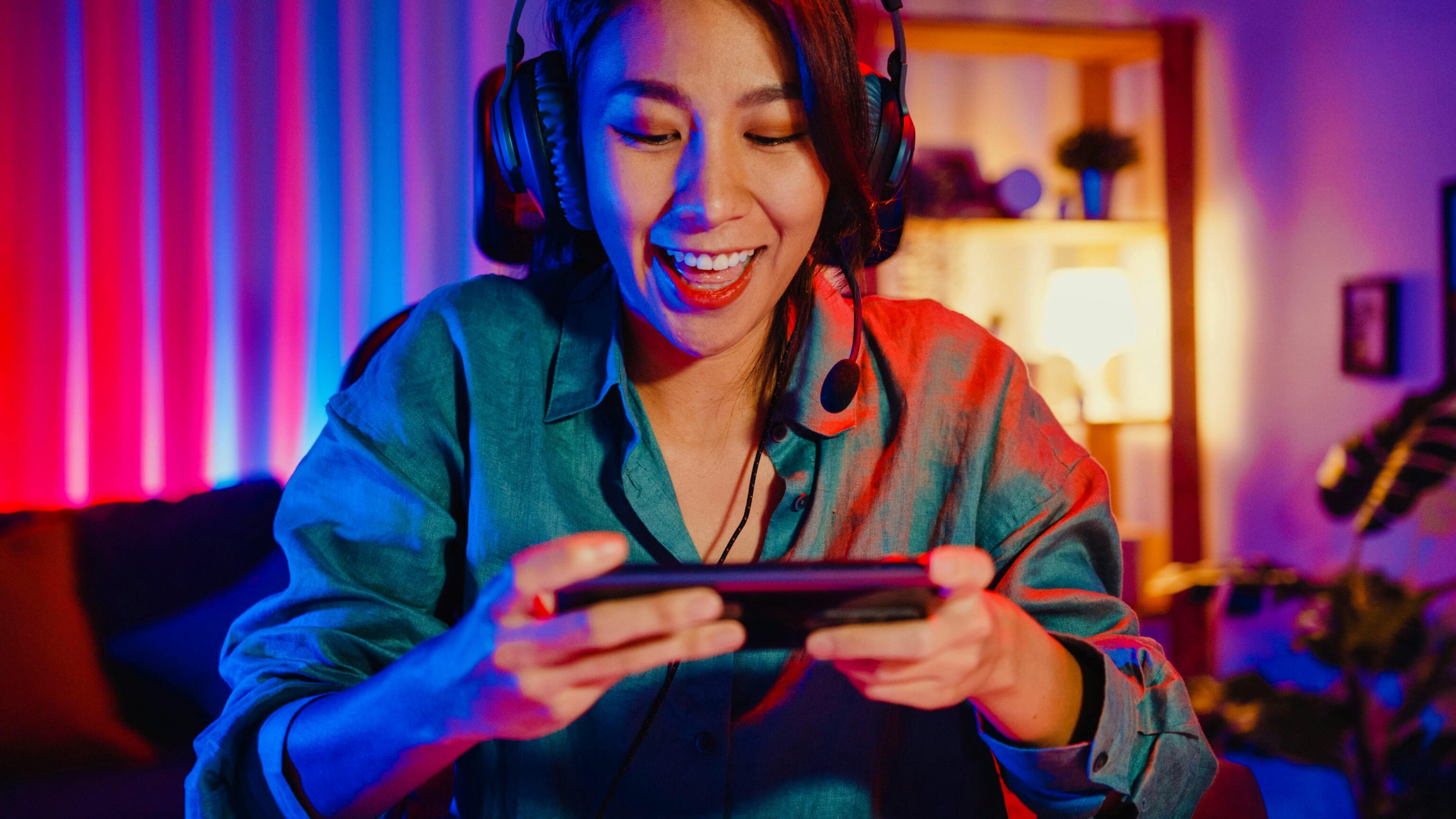 Donna con cuffie gioca con entusiasmo a un videogioco su cellulare in una stanza colorata.