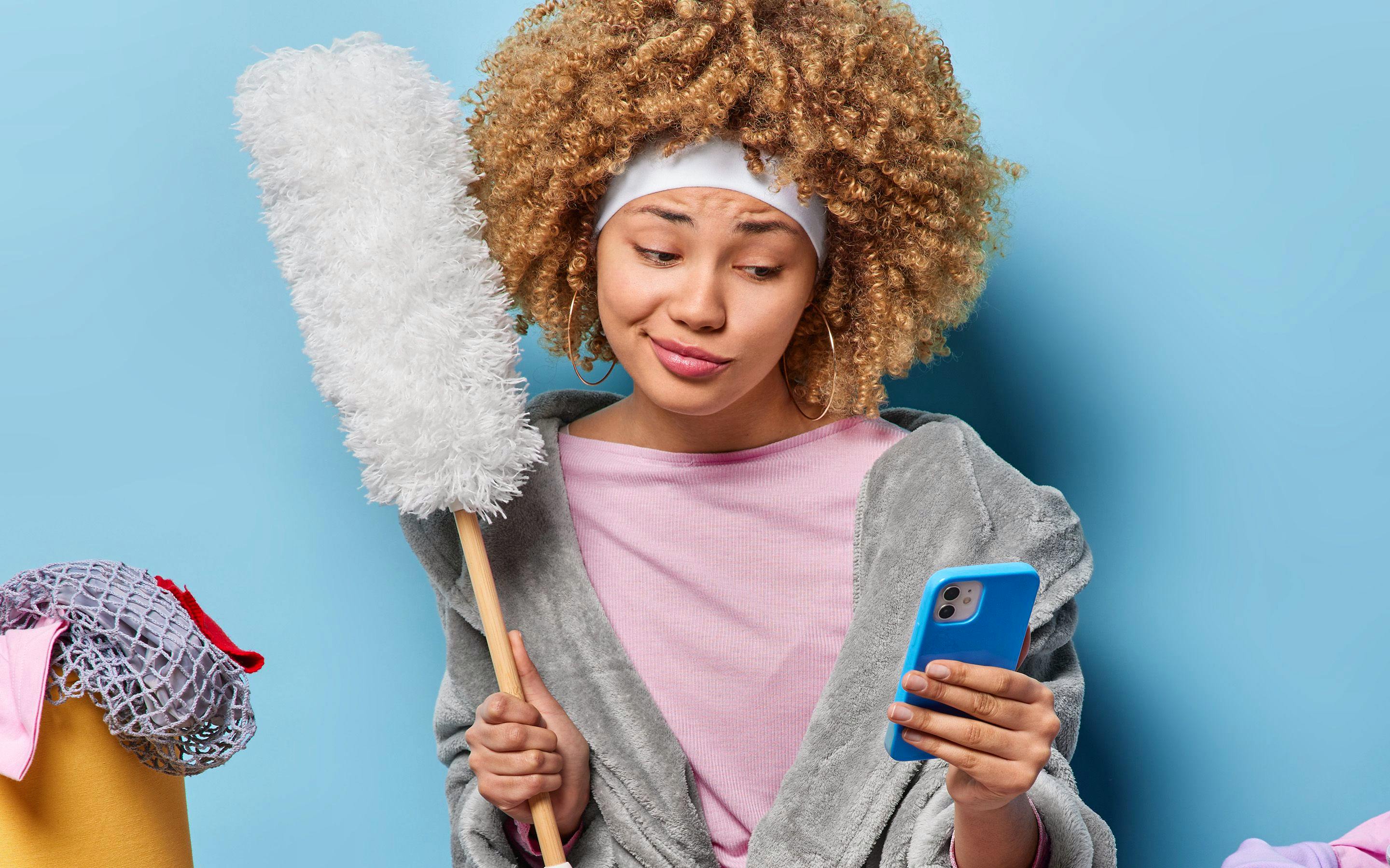 Una giovane donna tiene uno spolverino e guarda il telefono, distratta durante le pulizie.