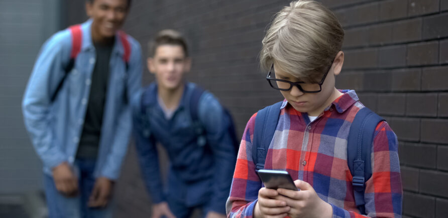 Ein Junge mit Brille und kariertem Hemd steht traurig mit einem Smartphone in der Hand, während zwei andere Jungen im Hintergrund lachen und ihn scheinbar verspotten.