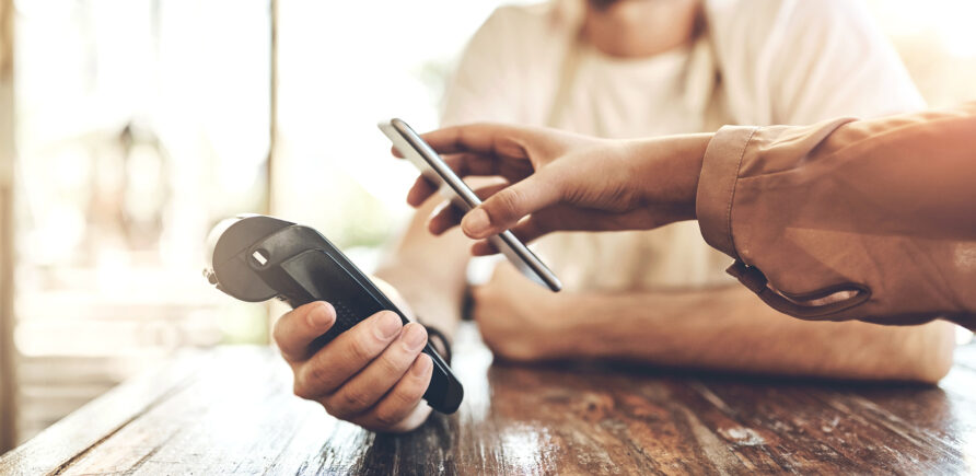 Ein Verkäufer hält ein Zahlterminal bereit. Eine Person bezahlt mit dem Smartphone.
