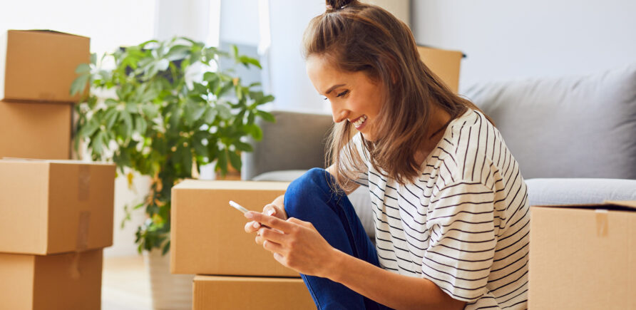 Una giovane donna è seduta tra scatole di trasloco e guarda il suo smartphone sorridendo - il trasloco è in corso.