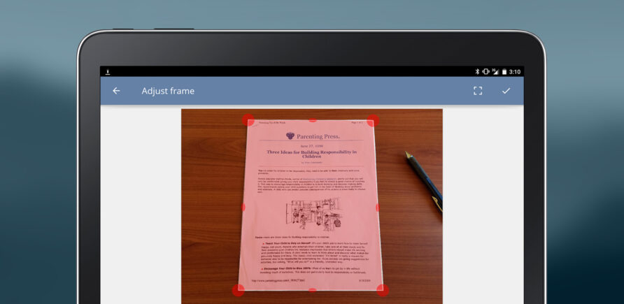 La capture d’écran de la tablette montre l’application de numérisation avec un document sur une table en bois et un cadre de découpe. Un stylo à bille se trouve à côté.