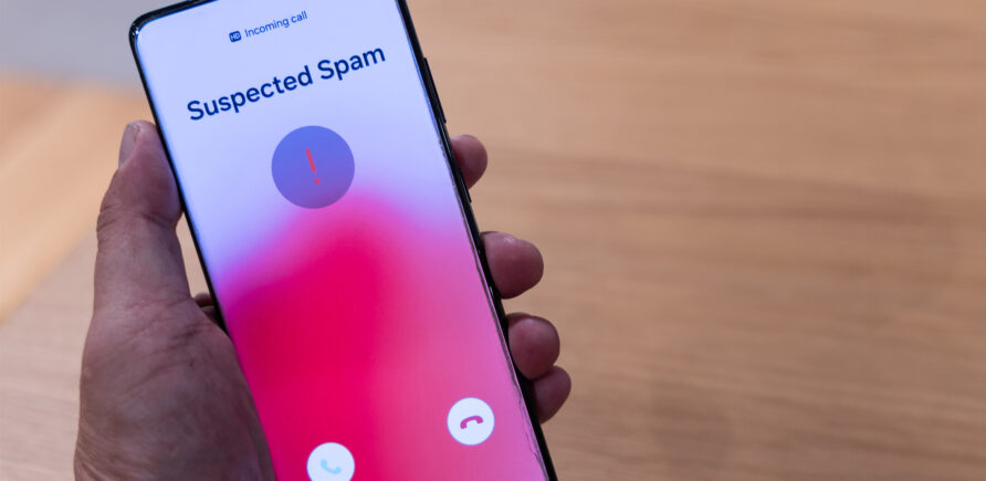 Une personne inconnue tient un téléphone portable dans ses mains. L'écran affiche un appel entrant intitulé 'Suspected Spam'.