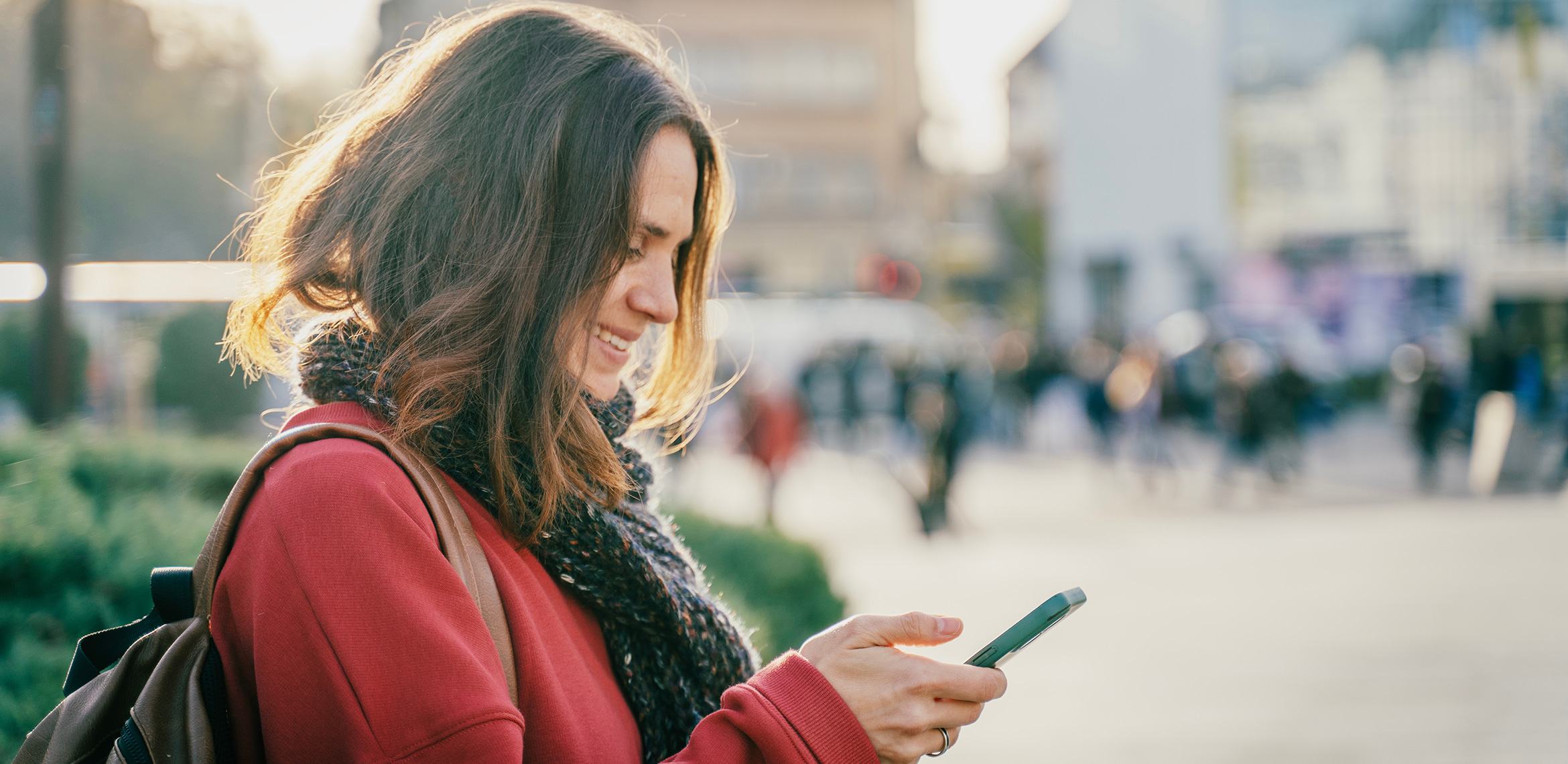 Eine Frau mit roter Jacke und Schal steht draussen auf einem sonnigen Platz in der Stadt. Sie lächelt und schaut auf ihr Smartphone.