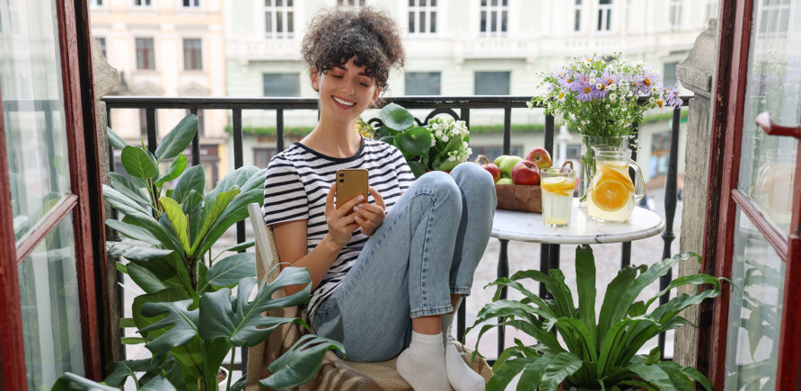 Eine junge Frau entspannt auf ihrem Balkon, umgeben von Pflanzen. Sie lächelt und schaut dabei auf ihr Smartphone. Auf dem Tisch steht ein erfrischendes Getränk bereit.