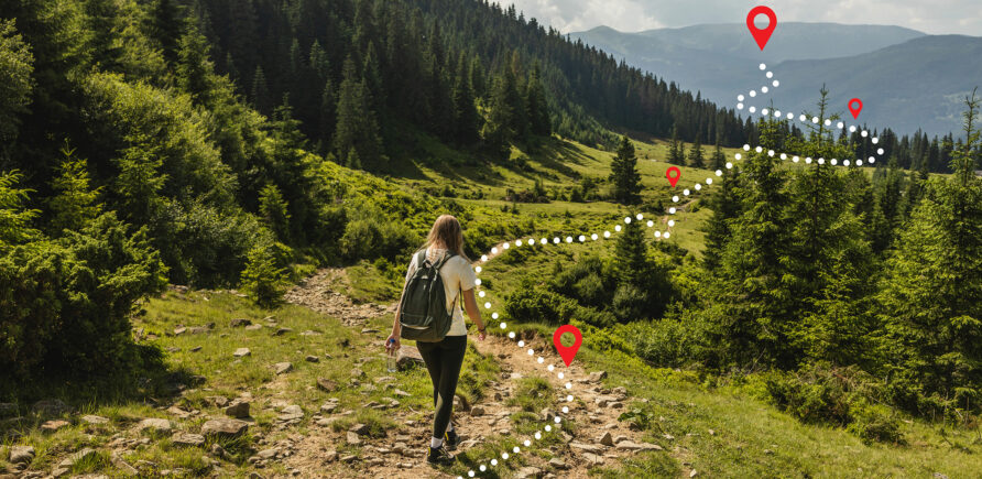 Eine Frau ist auf einem Wanderweg in den Bergen neben einem Wald unterwegs. Dabei hilft ihr eine digitale Anzeige.