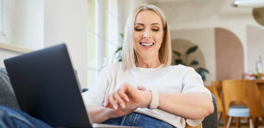 Une femme est assise sur un canapé dans son salon avec un ordinateur portable sur les genoux. Elle regarde sa montre connectée en souriant et touche l’écran.