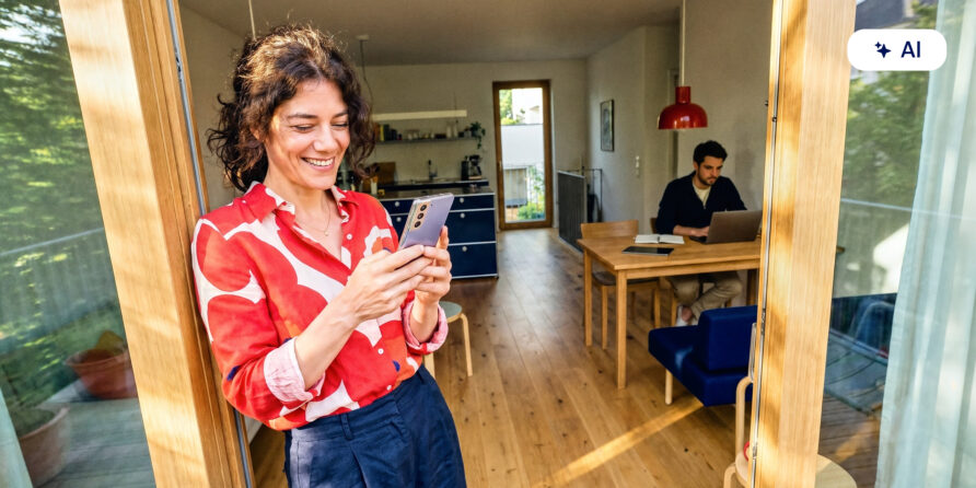 Eine Frau ist lächelnd am Smartphone im Eingang eines hellen, modernen Hauses, während im Hintergrund ein Mann am Notebook sitzt.